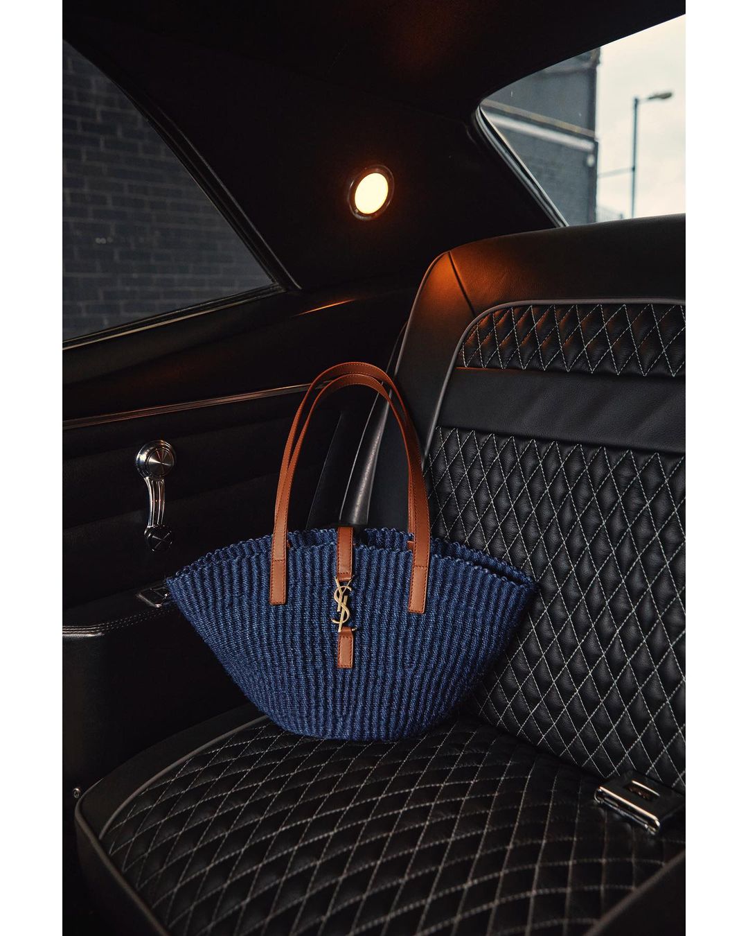 Saint Laurent bag on a quilted leather car seat with brown leather handles. The handbag is mostly navy and black with gold logo.