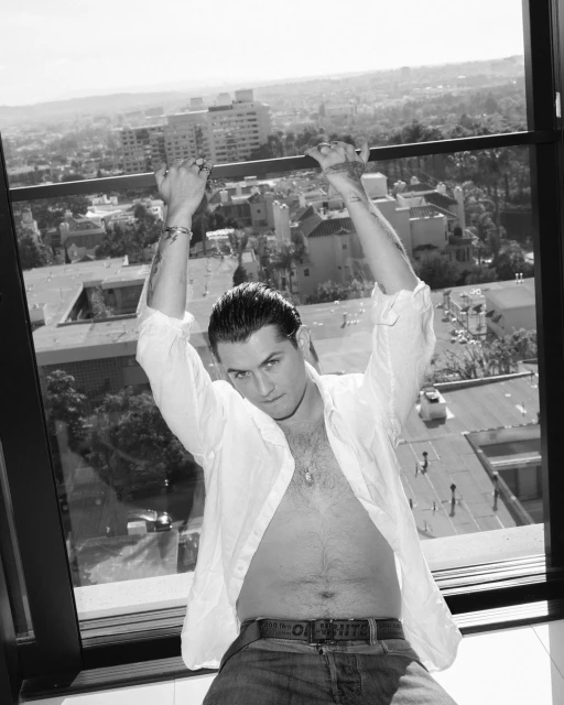 Black and white portrait of an adult male with an open white shirt and jeans, posing near a window with a cityscape background.