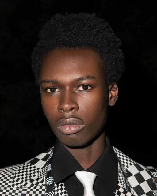 Portrait of a young African male in a checkerboard suit, black shirt, and white tie against a dark background, looking directly at the camera.