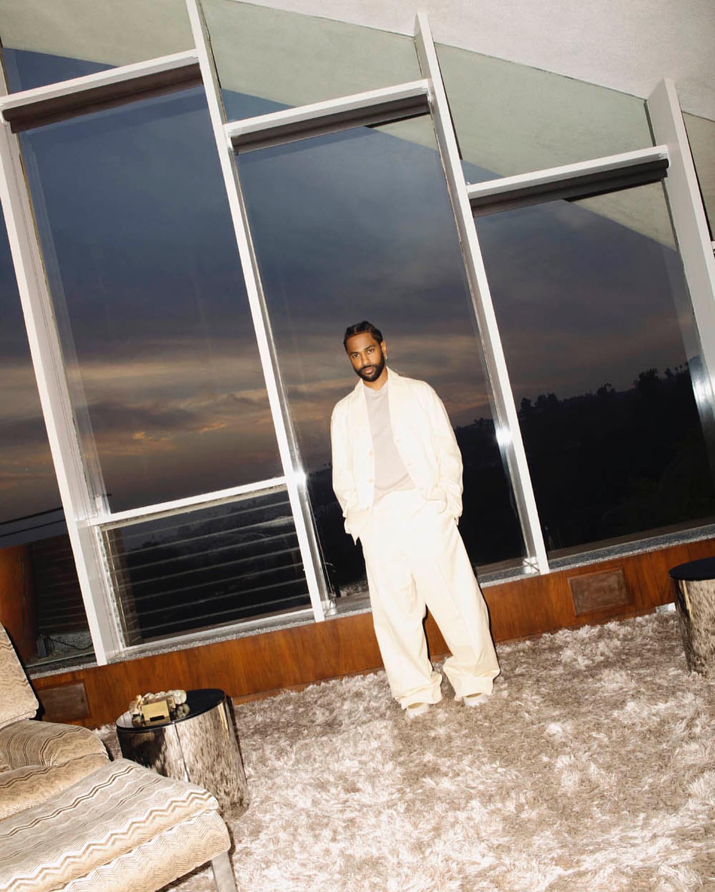 An adult male in a cream-colored suit poses in front of a large window. The soft, natural light highlights the textures of the carpet and furniture.