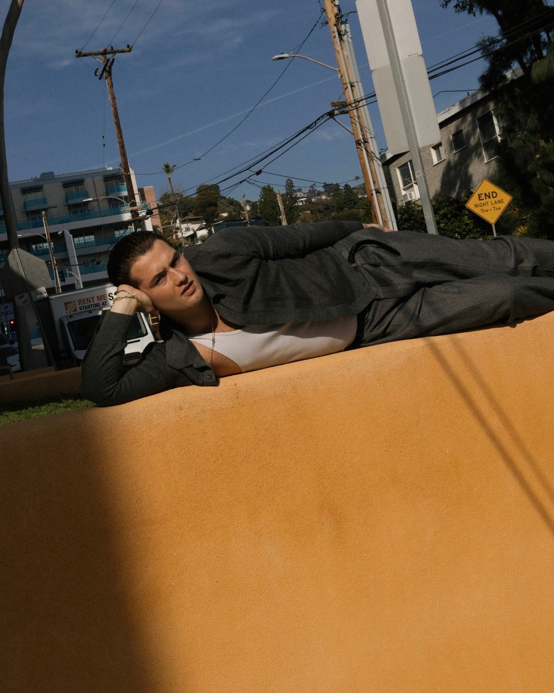 A young adult male in a suit lies on an orange wall with his head propped on his hand, posing in an outdoor urban setting.