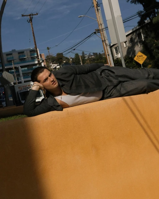 A young adult male in a suit lies on an orange wall with his head propped on his hand, posing in an outdoor urban setting.