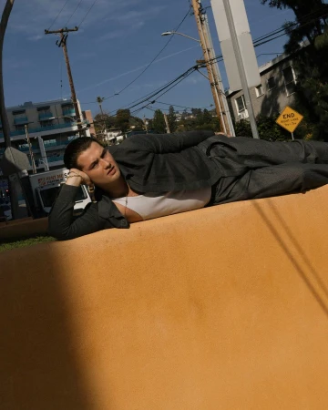 A young adult male in a suit lies on an orange wall with his head propped on his hand, posing in an outdoor urban setting.