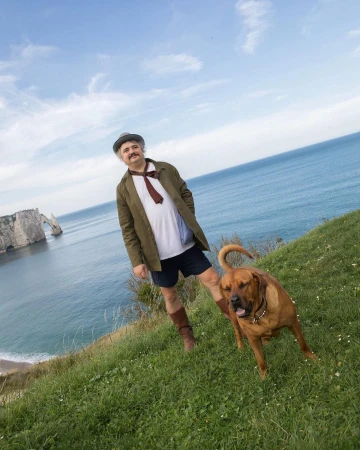 Adult male with a dog near a sea cliff. Man wears a hat and suit jacket. The dog is brown.  Ocean and white cliffs background.