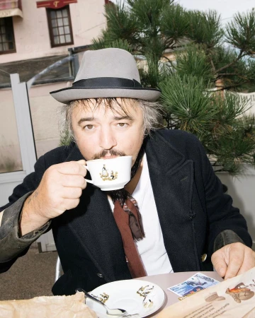 Man wearing grey fedora and black coat drinking from a patterned teacup outdoors by a pine tree