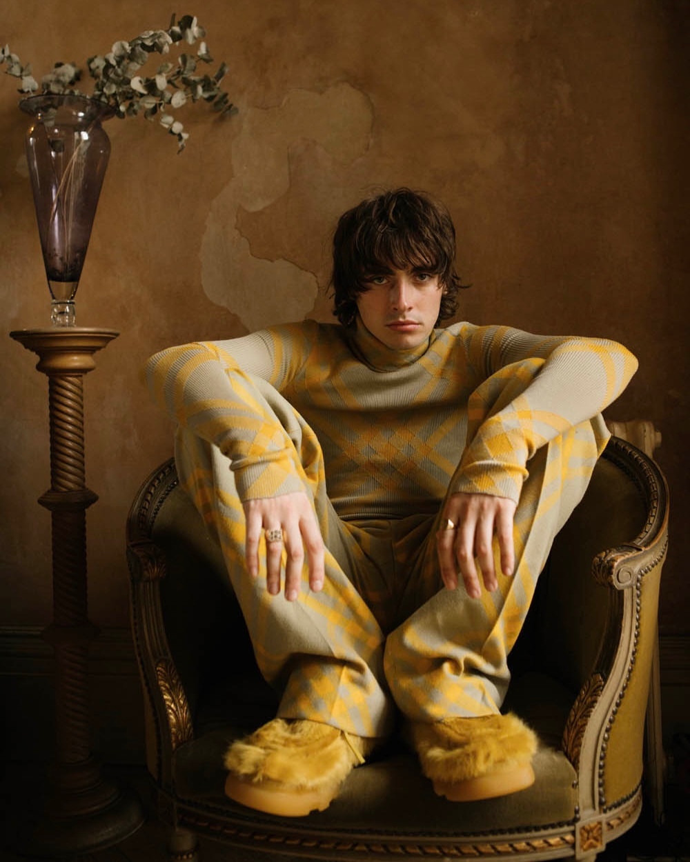 Stylish young adult in patterned sweater and pants sitting in a chair, accessorized with rings, flowers in vase behind.