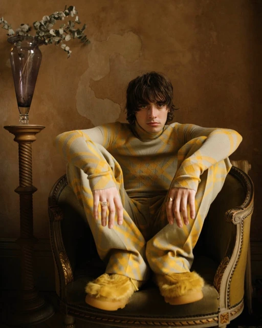 Stylish young adult in patterned sweater and pants sitting in a chair, accessorized with rings, flowers in vase behind.