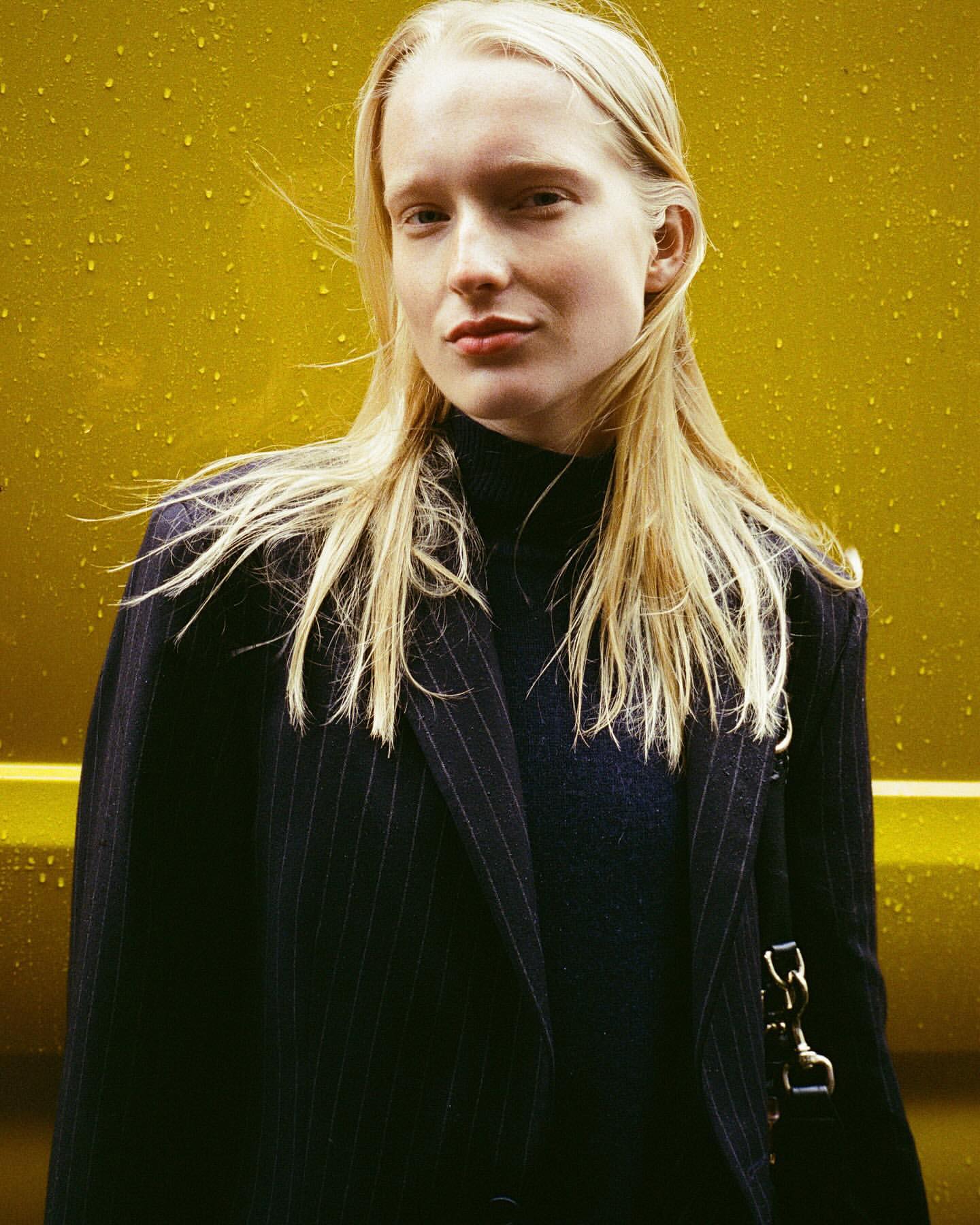 Blonde woman in pinstripe blazer stands against a yellow wall in a medium shot, fashion editorial, neutral expression, long blonde hair.