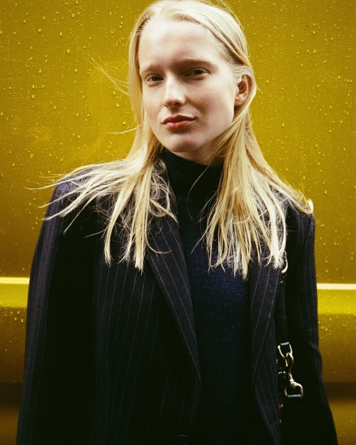 Blonde woman in pinstripe blazer stands against a yellow wall in a medium shot, fashion editorial, neutral expression, long blonde hair.