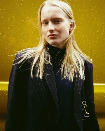 Blonde woman in pinstripe blazer stands against a yellow wall in a medium shot, fashion editorial, neutral expression, long blonde hair.