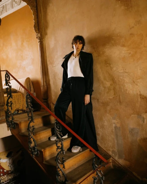 A young adult woman in a black coat and pants standing on a wooden staircase in a warm-toned building with architectural details.