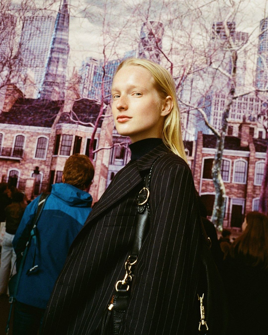 Fashion shot of a fair-skinned adult woman with blonde hair in a black pinstripe coat and black turtleneck, against a cityscape backdrop.