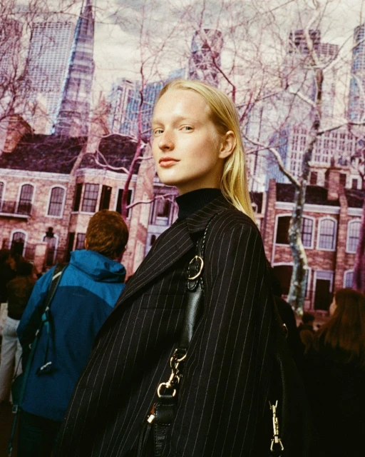 Fashion shot of a fair-skinned adult woman with blonde hair in a black pinstripe coat and black turtleneck, against a cityscape backdrop.