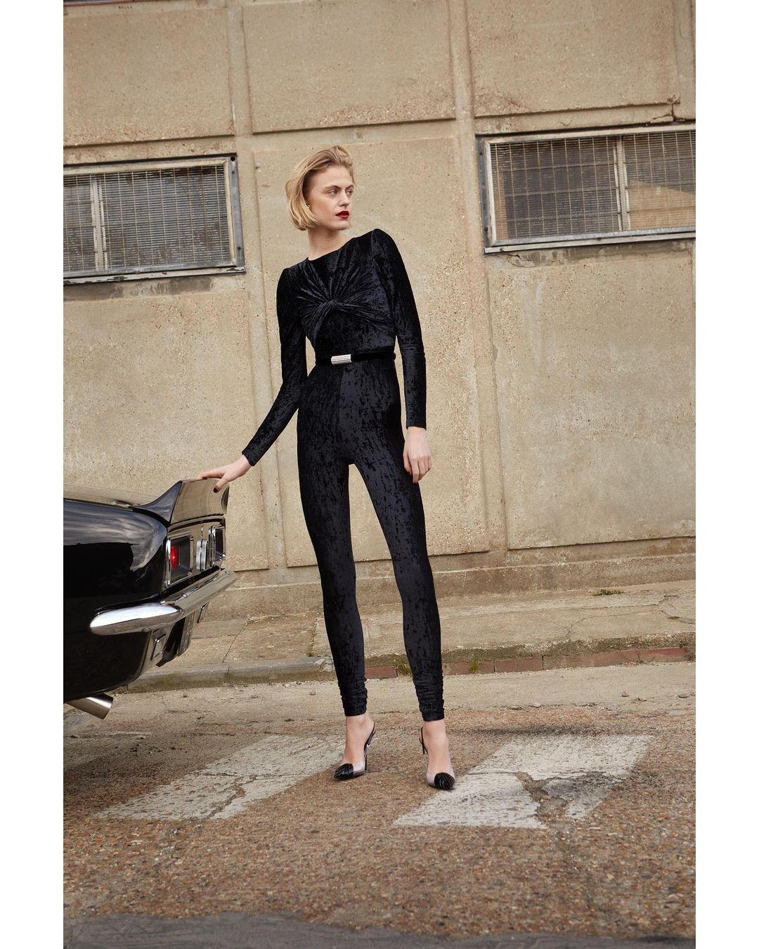 A slender woman with short blonde hair, dressed in a black velvet jumpsuit and heels, poses next to a black car in an urban setting.