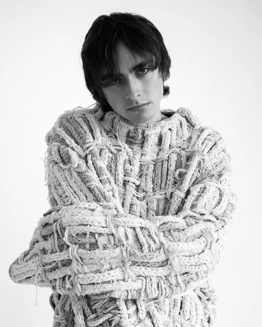 Monochrome portrait of a young adult posing indoors with arms crossed, wearing a hand-knitted textured wool sweater against a white background.