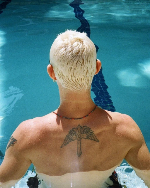 Back view of a shirtless adult in a pool with short blond hair, ear piercings, beaded necklace and a large angel wing tattoo.