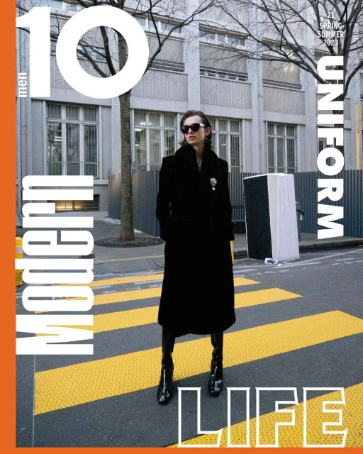 Magazine cover featuring a person in a long black coat and patent boots, posing on a yellow crosswalk with a building backdrop.