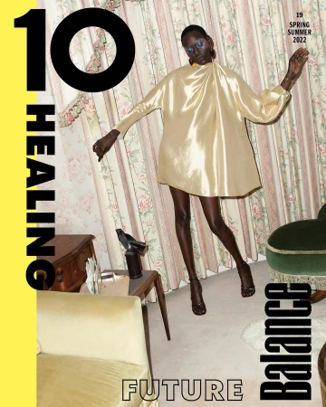 Black woman wearing a glossy gold satin minidress and strappy heels standing in a vintage living room with floral curtains