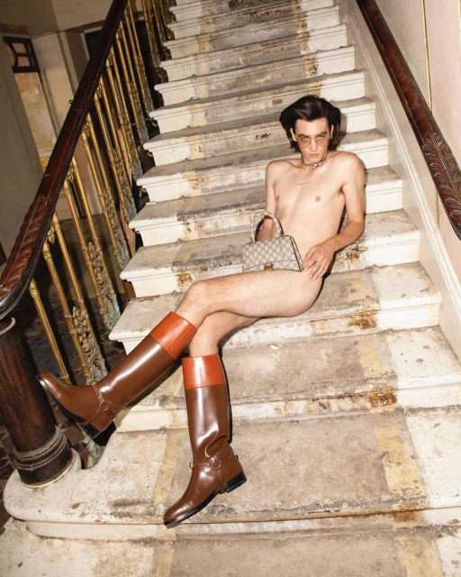 A shirtless man poses on a staircase wearing riding boots and holding a handbag, indoor setting with golden light.