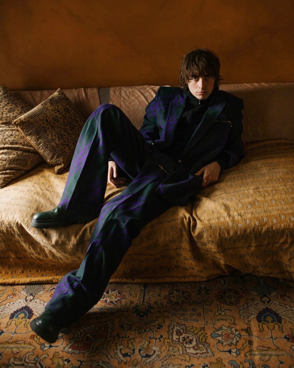 Person in a plaid suit lying on a bed with pillows, showcasing a relaxed pose in an indoor setting with warm, diffused lighting.