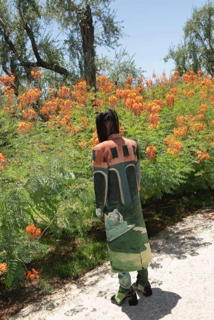Adult standing among orange flowering bushes outdoors wearing a long coat and light green pants and shoes in natural light.