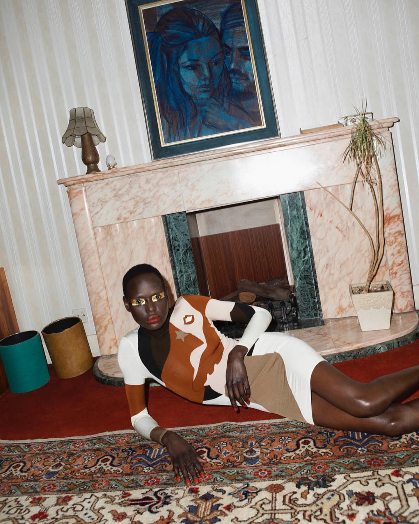 An African woman with golden eyeshadow and a patterned dress poses in a room with a fireplace, painting, and ornate carpet.