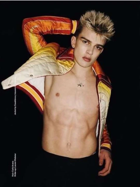 Portrait of a young adult male with blond spiky hair, wearing an unbuttoned orange and white racing jacket, against a black background.