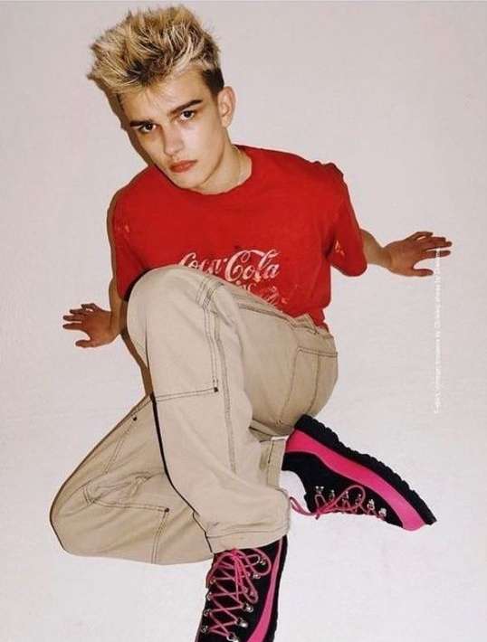 An adult male with bleached blond hair, wearing a red Coca-Cola T-shirt and khaki pants, poses indoors with pink and black hiking boots.