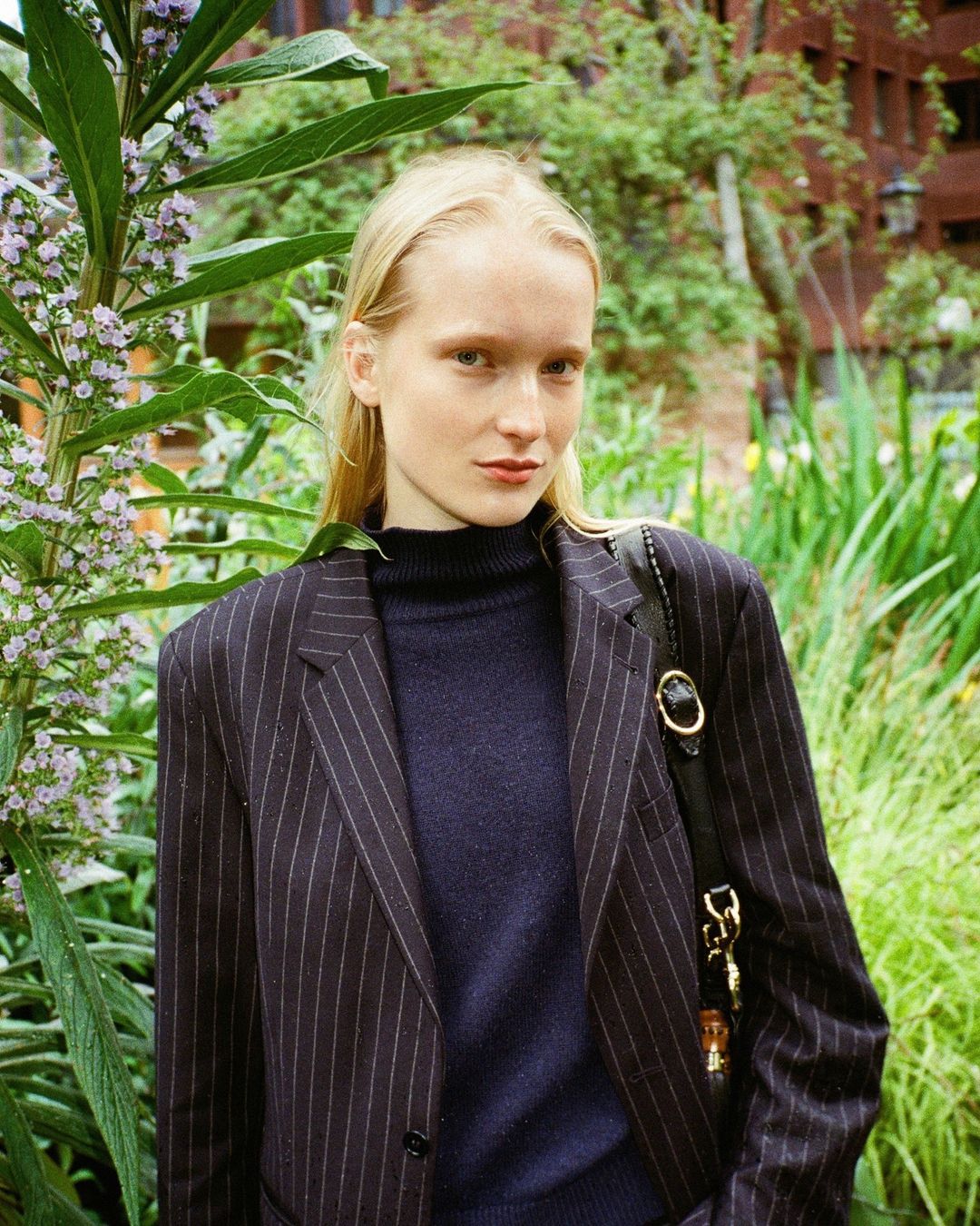 Portrait of a fair-skinned woman with blonde hair in pinstripe blazer, standing outdoors amidst lush greenery and foliage.