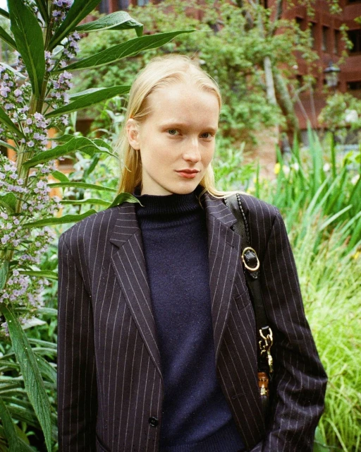 Portrait of a fair-skinned woman with blonde hair in pinstripe blazer, standing outdoors amidst lush greenery and foliage.