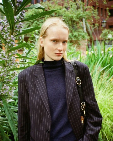 Portrait of a fair-skinned woman with blonde hair in pinstripe blazer, standing outdoors amidst lush greenery and foliage.