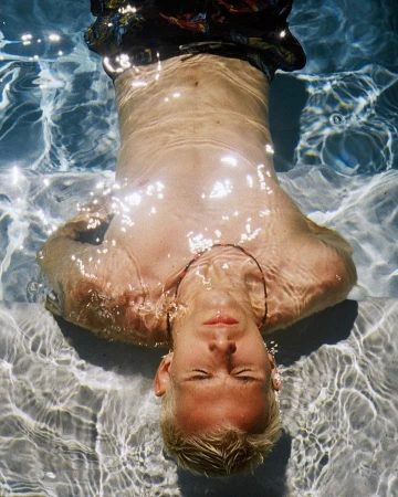 Underwater portrait of a fair-skinned adult male with blond hair floating in a pool, showcasing water ripples and bright, natural lighting.