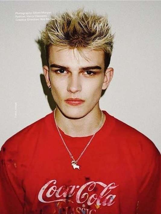 Close-up portrait of a young adult male with blonde hair, wearing a Coca-Cola logo t-shirt and silver chain with charm against plain background.