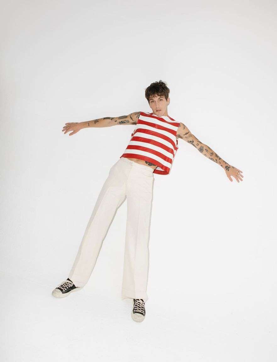 Young adult with tattoos poses on a white background, wearing a red and white striped tank top, cream pants, and patterned sneakers.