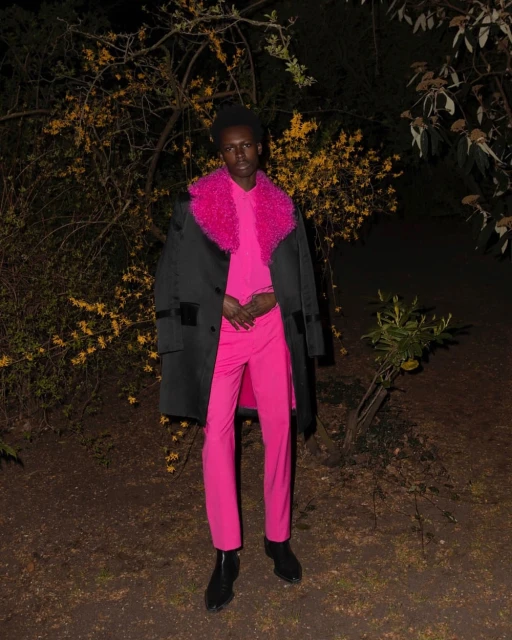 Adult male wearing a bright pink suit with a furry collar, standing in a dimly lit outdoor environment with foliage.