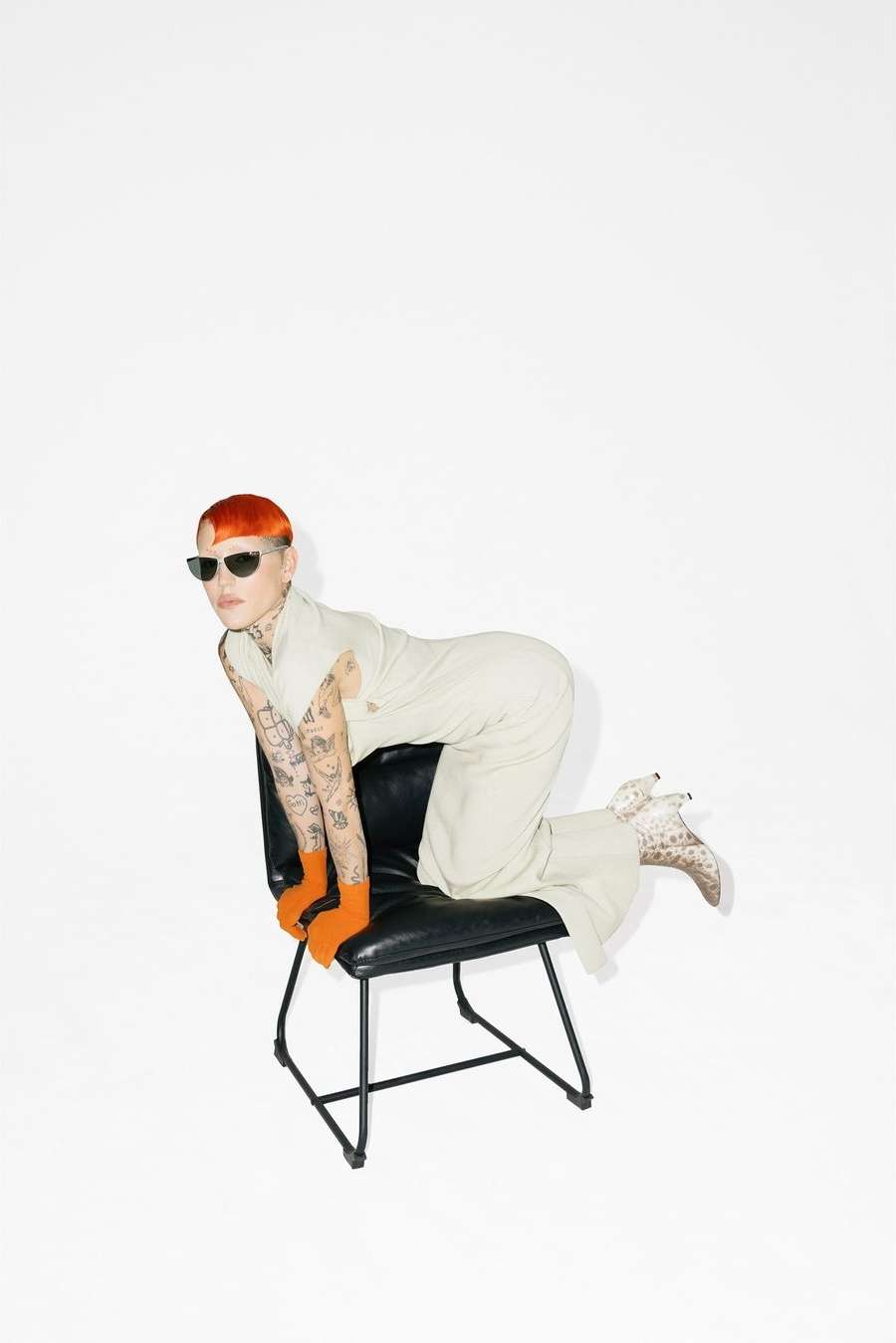 Woman with orange hair and sunglasses in a sleeveless top and pants kneeling on a black chair in front of a white backdrop.
