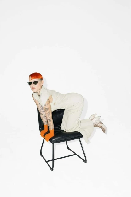 Woman with orange hair and sunglasses in a sleeveless top and pants kneeling on a black chair in front of a white backdrop.