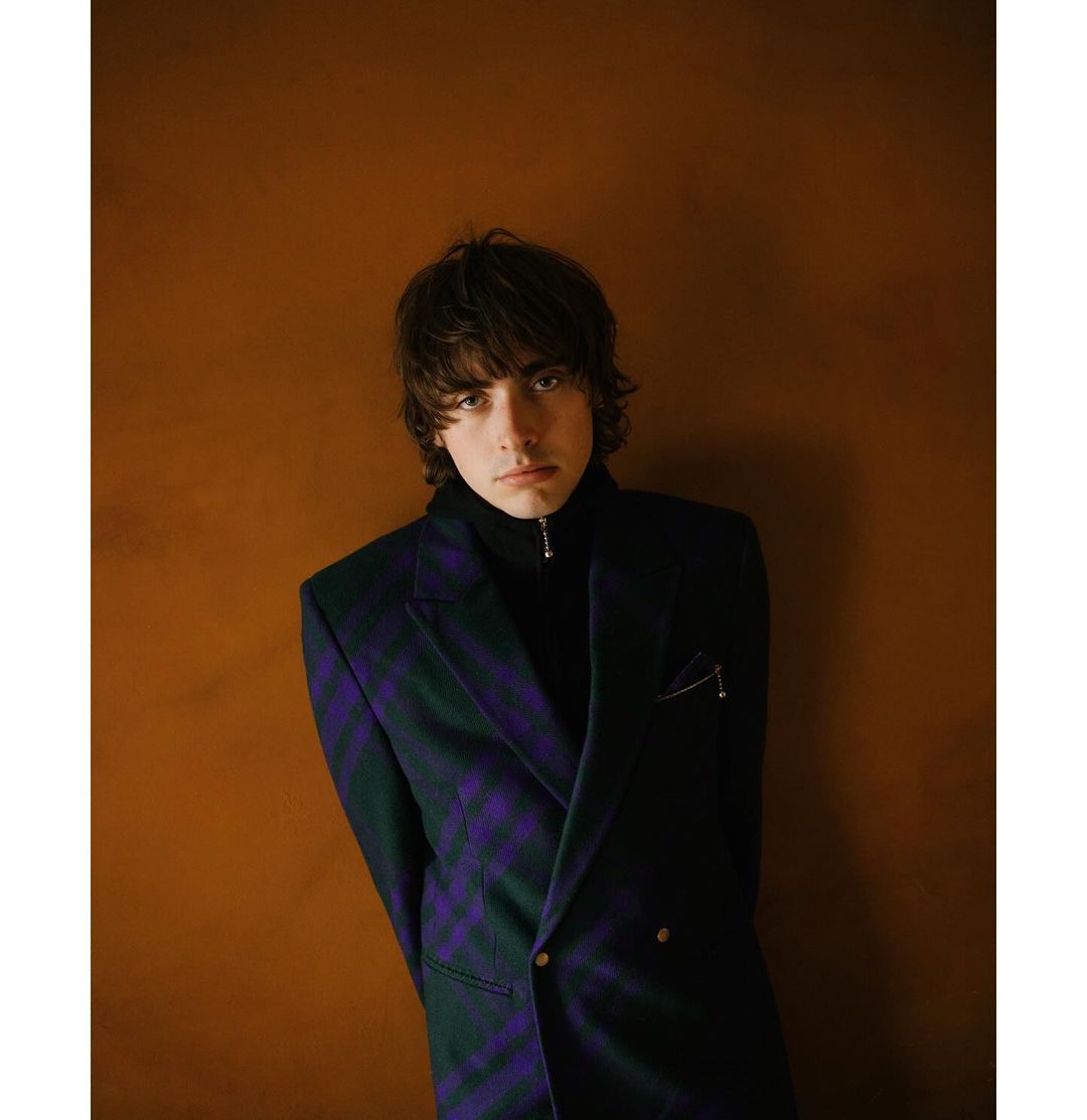 Portrait of a young adult man with dark hair wearing a patterned blazer, captured against a solid brown background in an indoor setting.