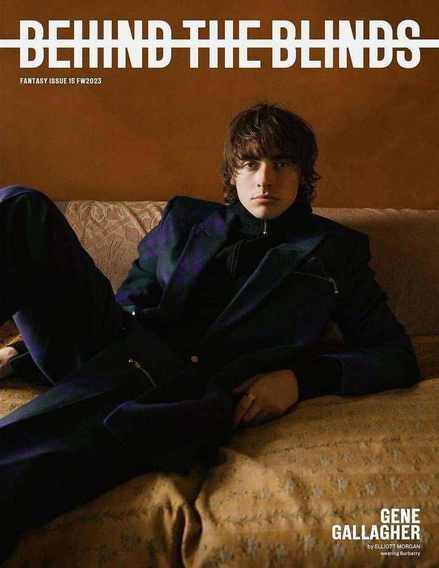 A young man wearing a dark plaid blazer and trousers reclines on a textured sofa against a warm terracotta wall.