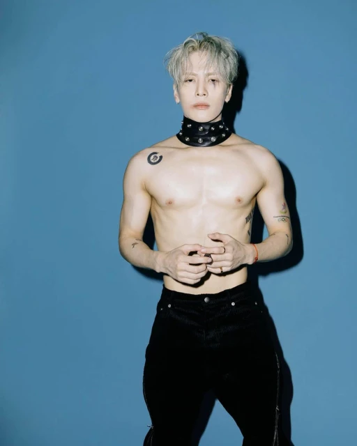 Shirtless man with silver hair and black pants poses against a solid blue background wearing a studded choker necklace.