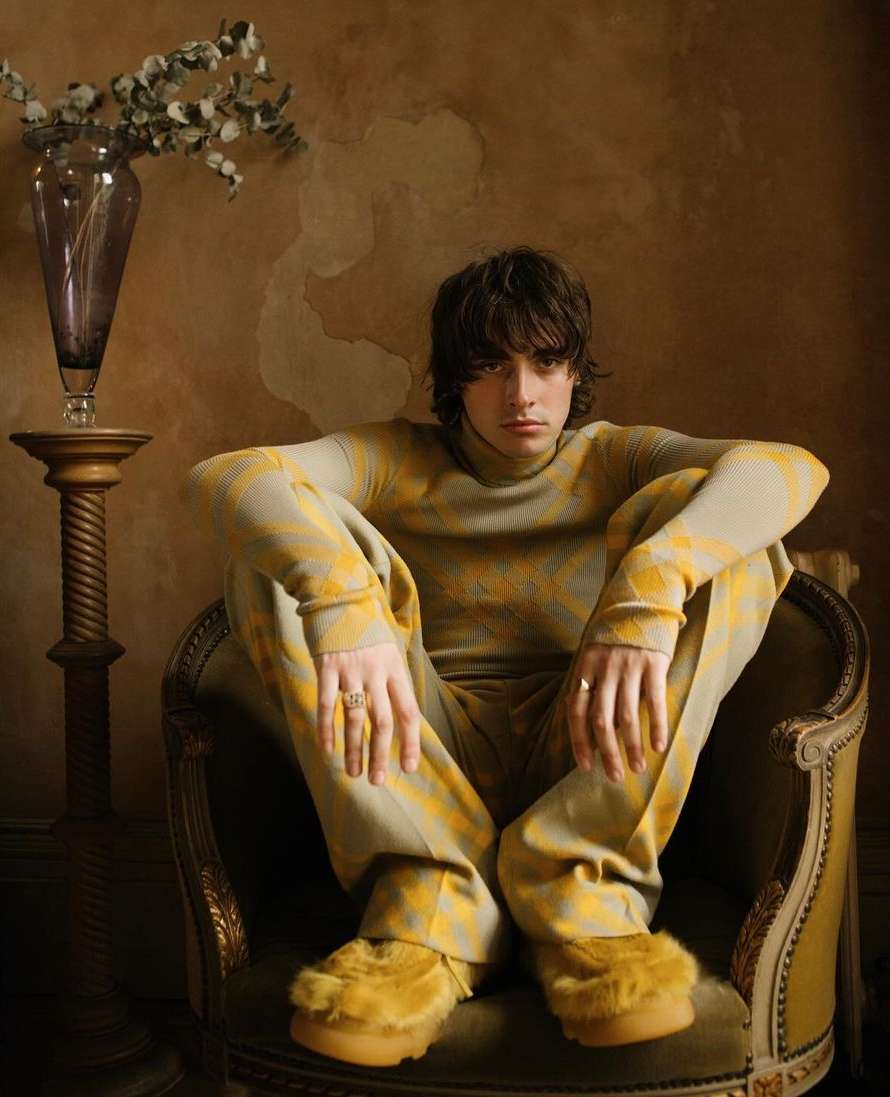 A young adult with dark hair sits in a vintage chair wearing a tan and yellow patterned outfit with furry yellow platform shoes in a room.