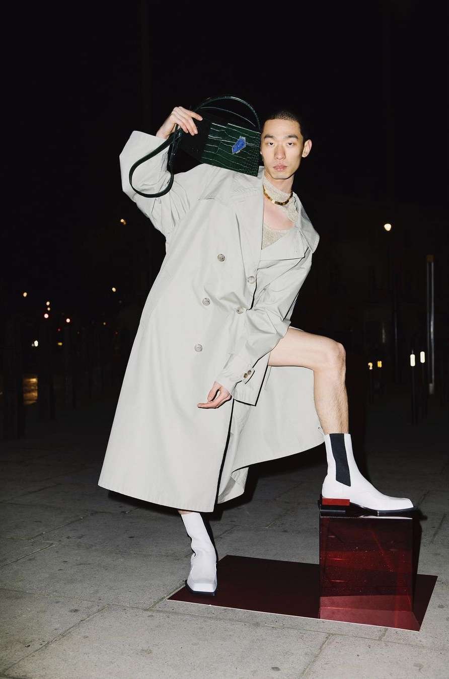 Asian model poses outdoors at night in trench coat and white boots, holding a box bag, with cityscape lights blurred in background.