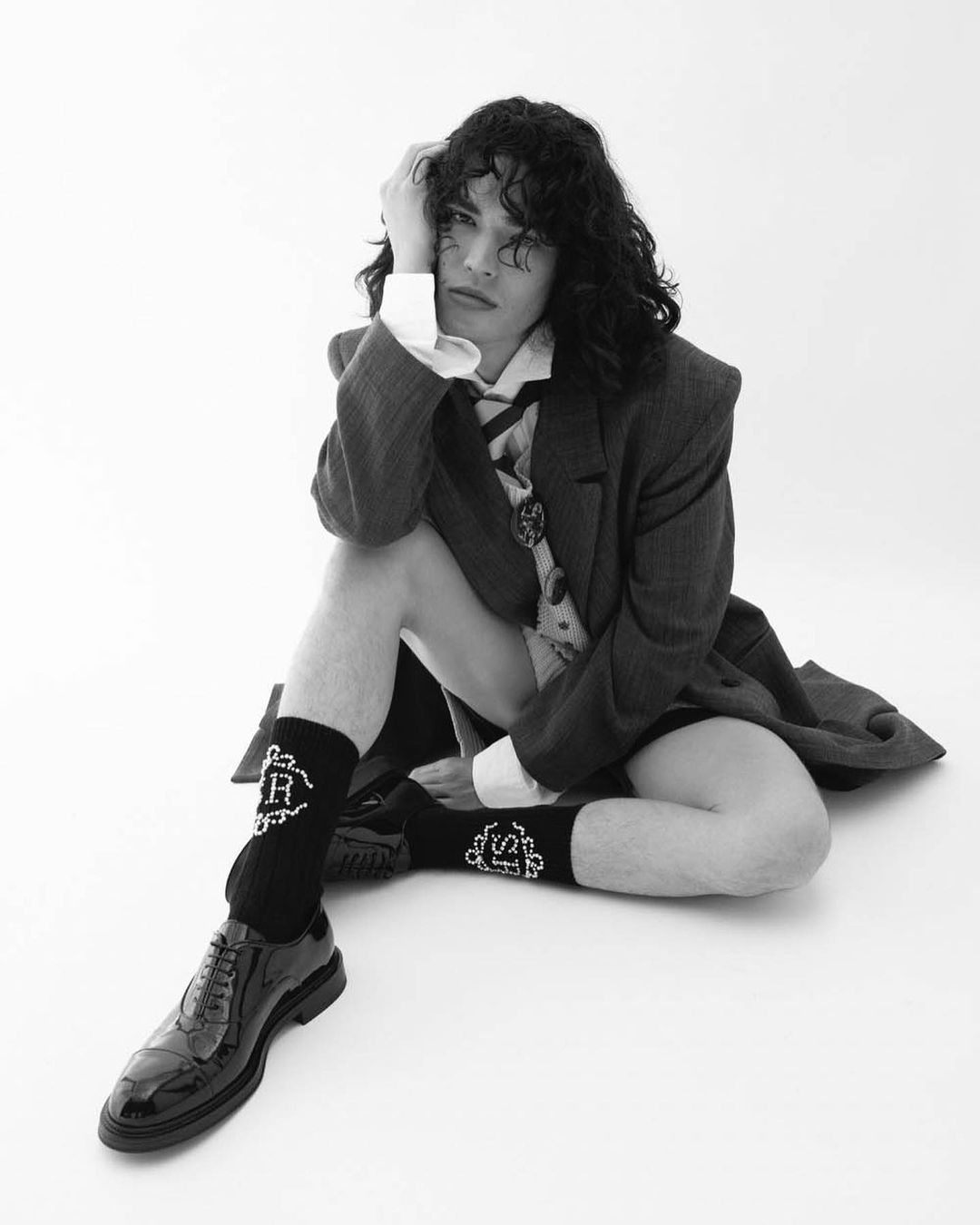 Monochrome portrait of a man sitting on the floor wearing an oversized blazer, white shirt, patterned tie, black socks, and dress shoes.