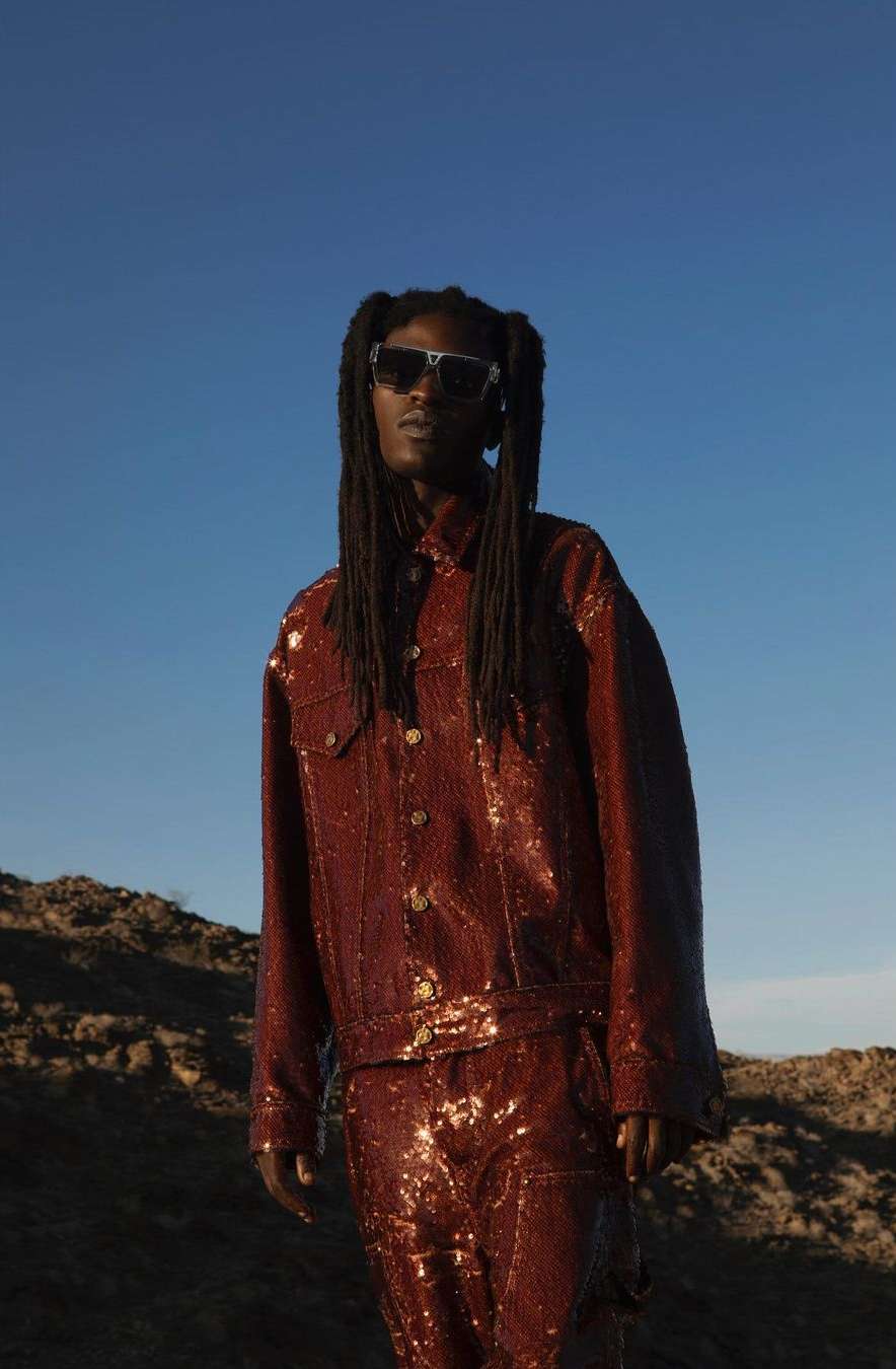 Black man with long dreadlocks wearing dark sunglasses and a glossy dark red textured jacket against a clear blue sky and rocky landscape