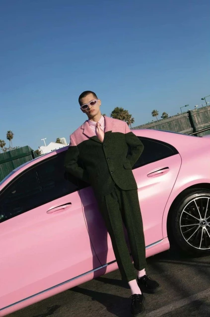 A man in a two-tone suit leans against a pink car with palm trees and blue sky in the background in an outdoor fashion shoot.