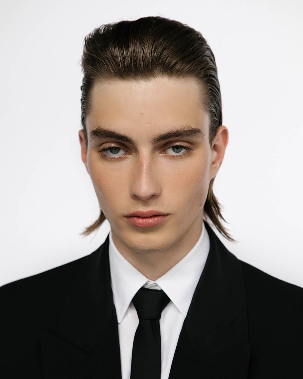Portrait of a man with slicked-back hair wearing a black suit and tie against a white background by Elliott Morgan