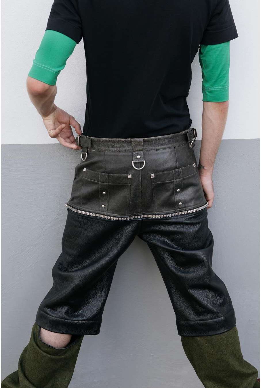 Mid-shot of an adult wearing a black t-shirt and dark leather shorts-style trousers, posing in front of a two-tone grey wall.