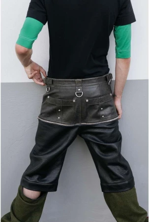 Mid-shot of an adult wearing a black t-shirt and dark leather shorts-style trousers, posing in front of a two-tone grey wall.