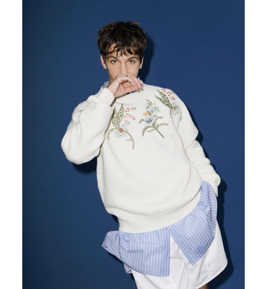 Portrait of a young adult male posing in a cream sweater with floral embroidery, paired with a blue-striped shirt and white shorts, against a navy backdrop.
