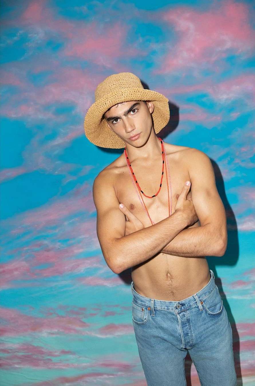 Young adult male in straw hat and jeans poses shirtless against a vibrant blue and pink cloud-patterned backdrop in a medium shot.